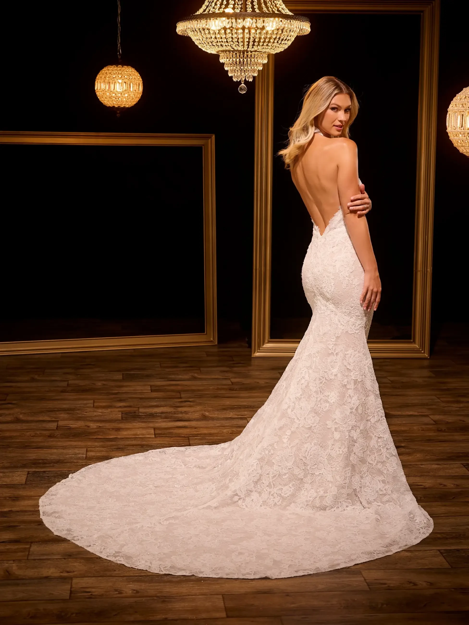 A woman in a backless white wedding dress with a long train, standing against a dark background with decorative chandeliers and gold-framed mirrors.