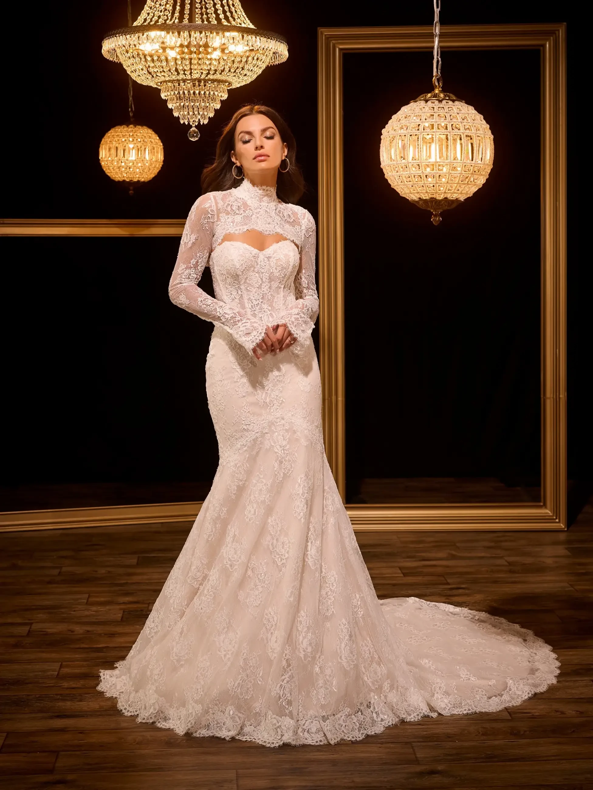 A woman in a elegant white lace wedding dress stands in a dimly lit room adorned with chandeliers, showcasing a fitted silhouette and long sleeves.