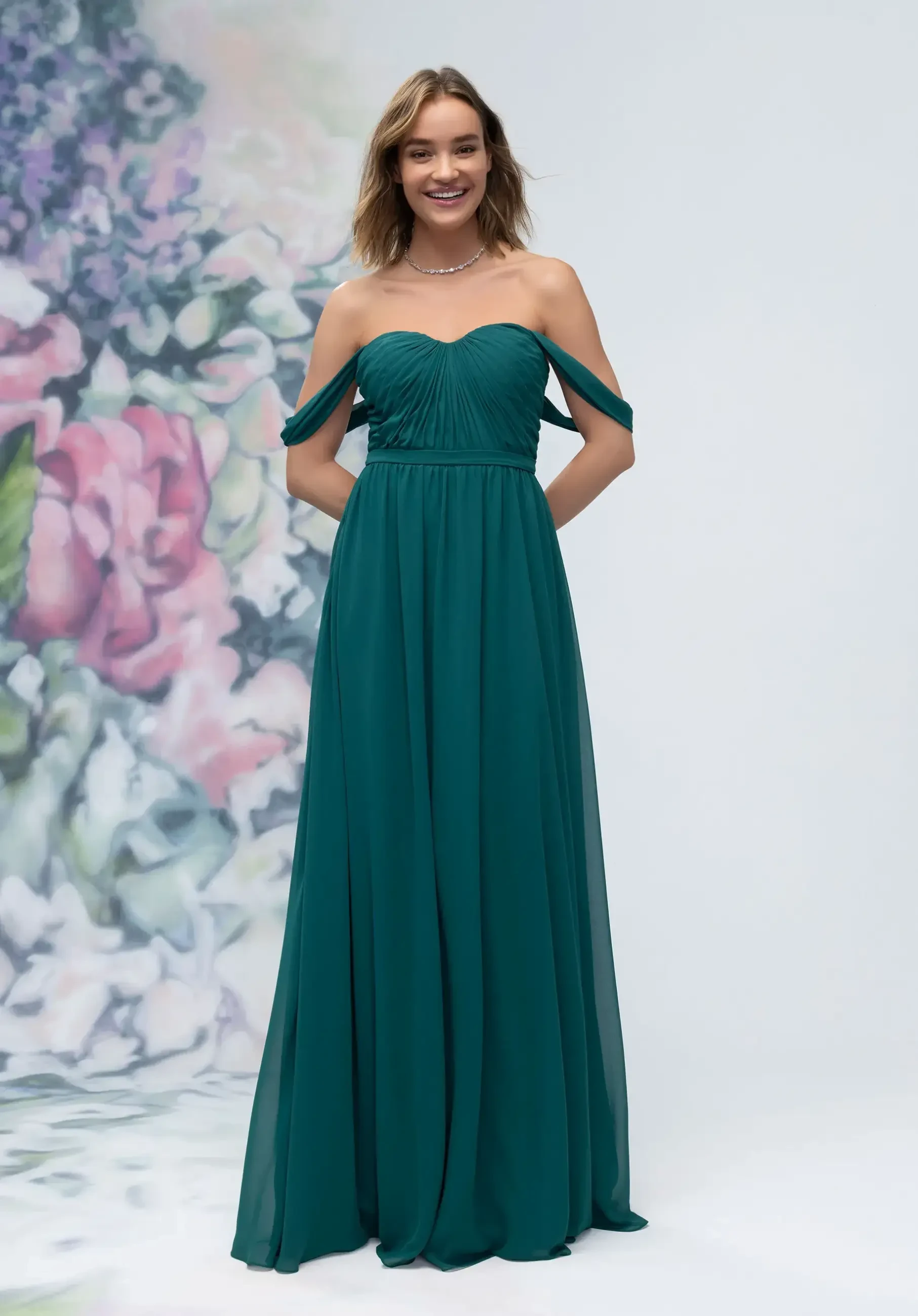 A woman in a teal, off-the-shoulder gown stands smiling against a blurred floral backdrop. The dress has a flowing skirt, conveying elegance and joy.