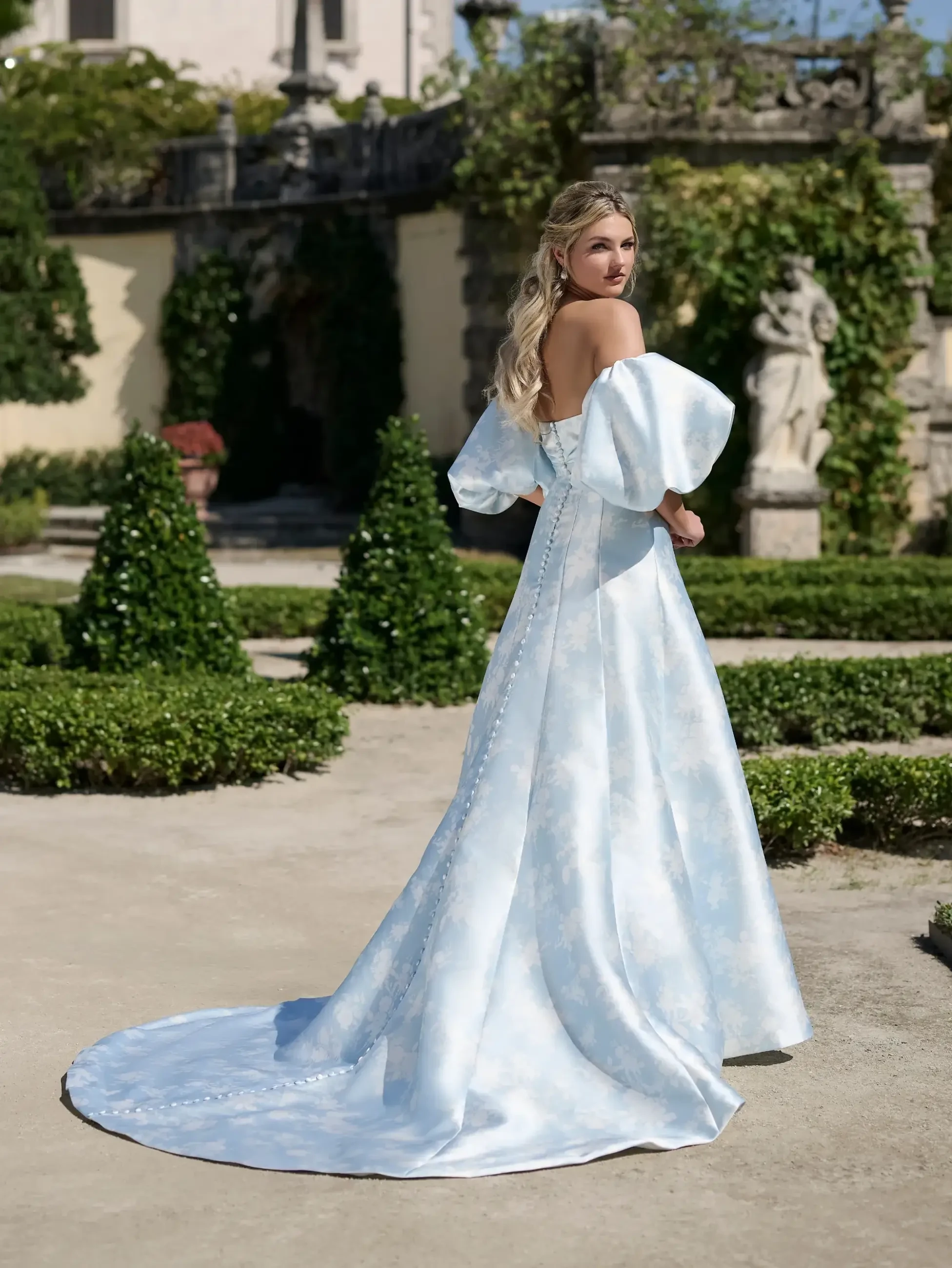 A woman wearing an elegant light blue gown with puffed sleeves stands outside in a garden, facing away from the camera. The gown has a long train and is surrounded by manicured hedges and greenery.