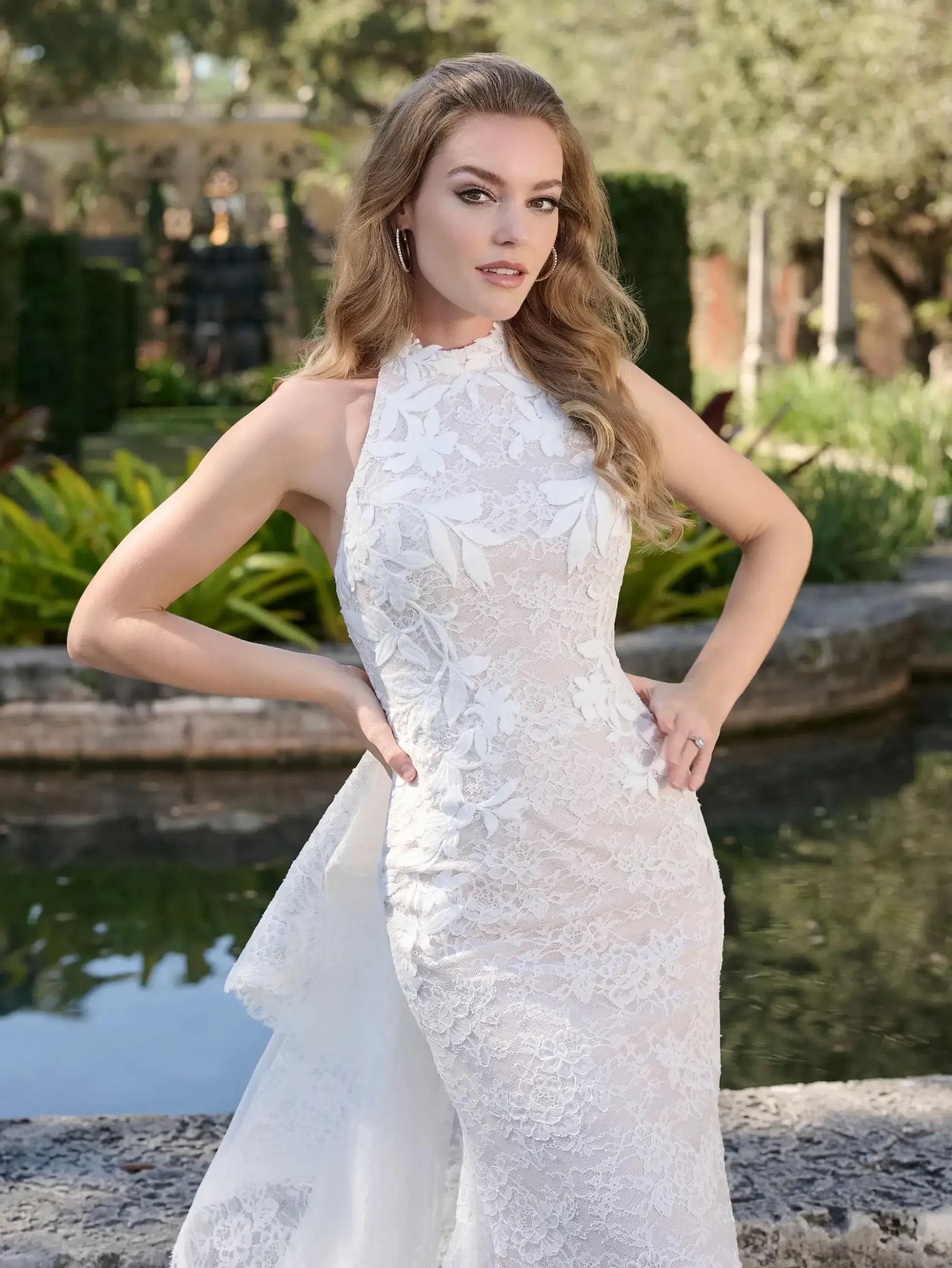 A woman wearing a white lace gown poses confidently in front of a garden and water feature.