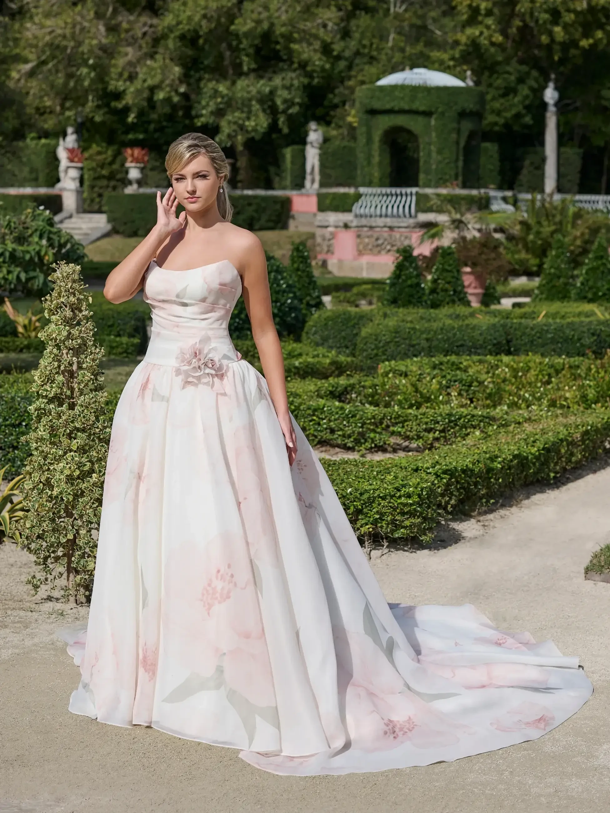 A woman in a strapless floral gown stands in a lush garden with manicured hedges and statues.