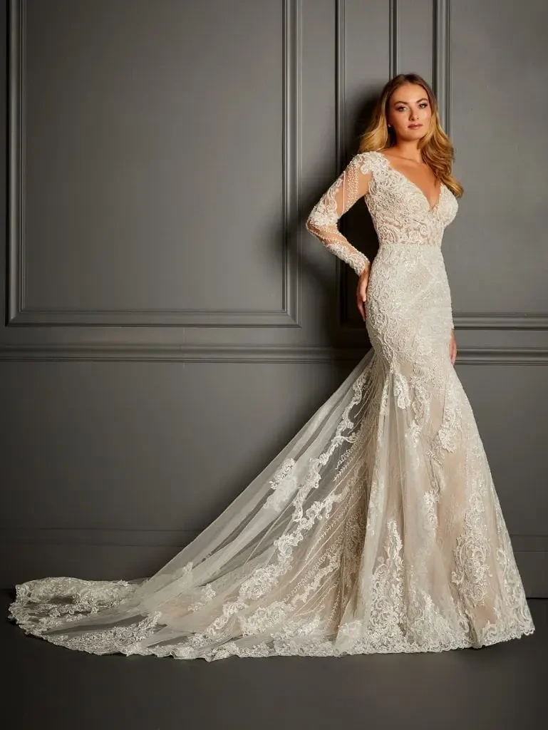 A woman in a lace wedding dress poses against a gray paneled wall. The dress features a long train, intricate lace details, and a deep V-neck, exuding elegance.