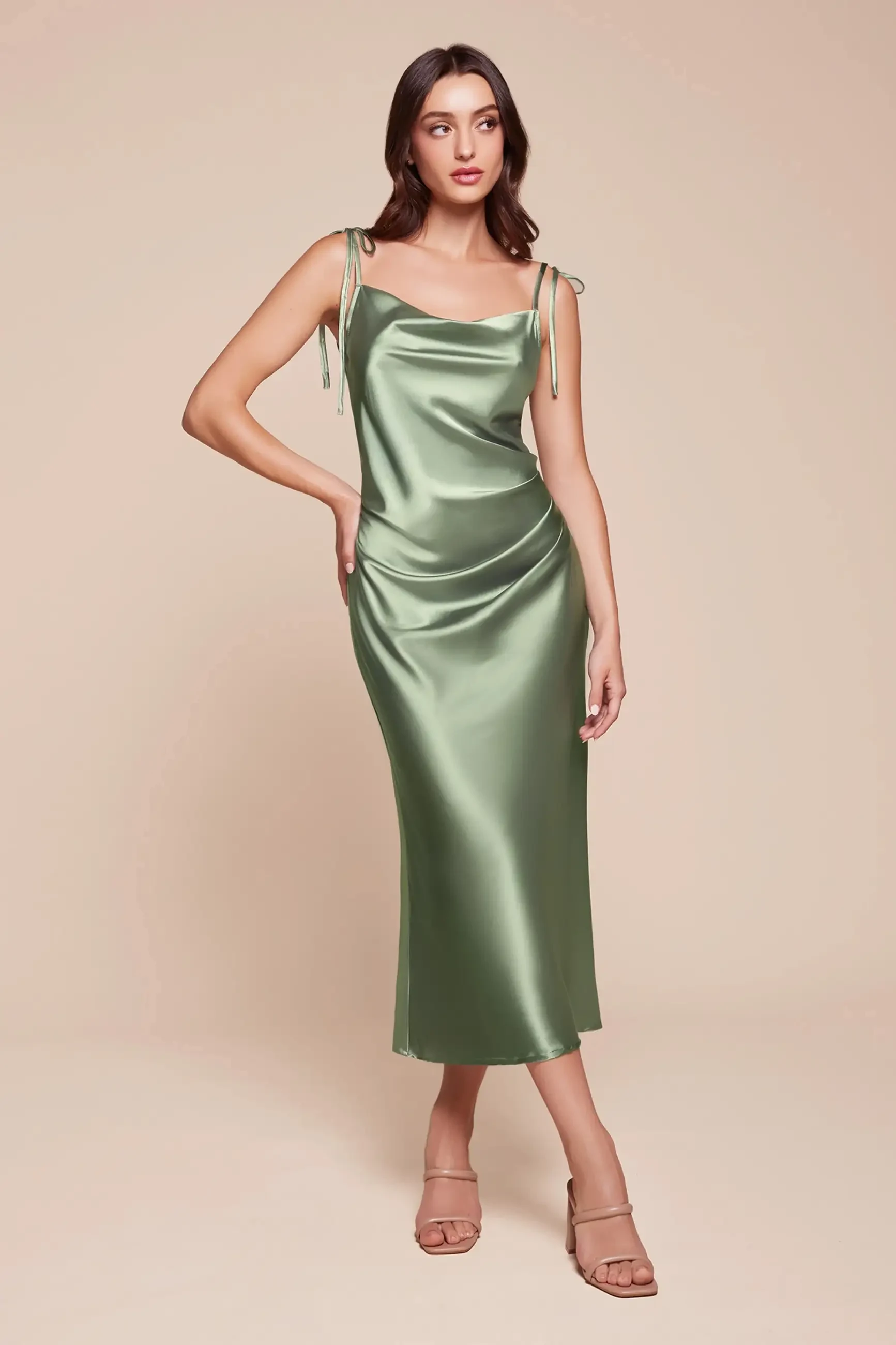 Woman wearing a sleek, satin sage green midi dress with tied straps, standing confidently against a beige background. She wears beige sandals, conveying elegance.