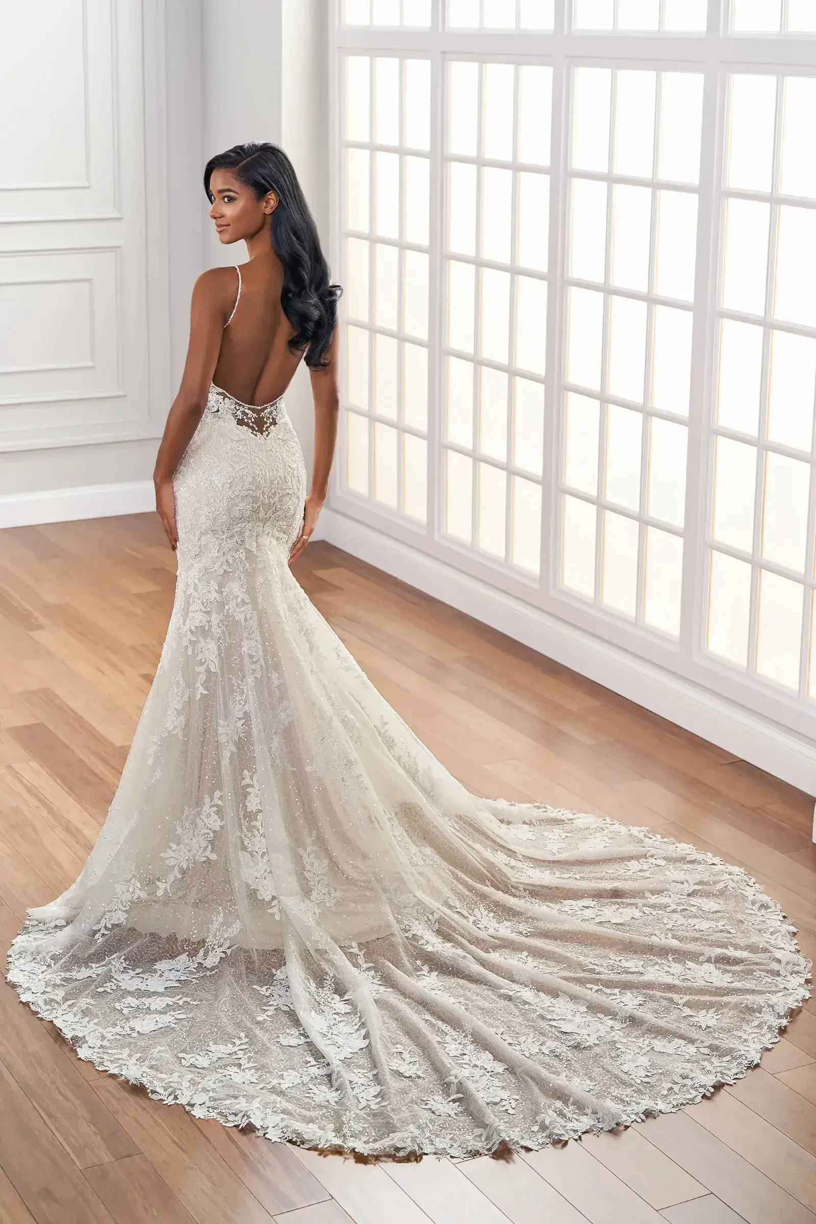 Model is wearing a sparkling fit and flare gown with beaded lace appliqué, a deep plunge neckline, beaded spaghetti straps, and an 86-inch train.