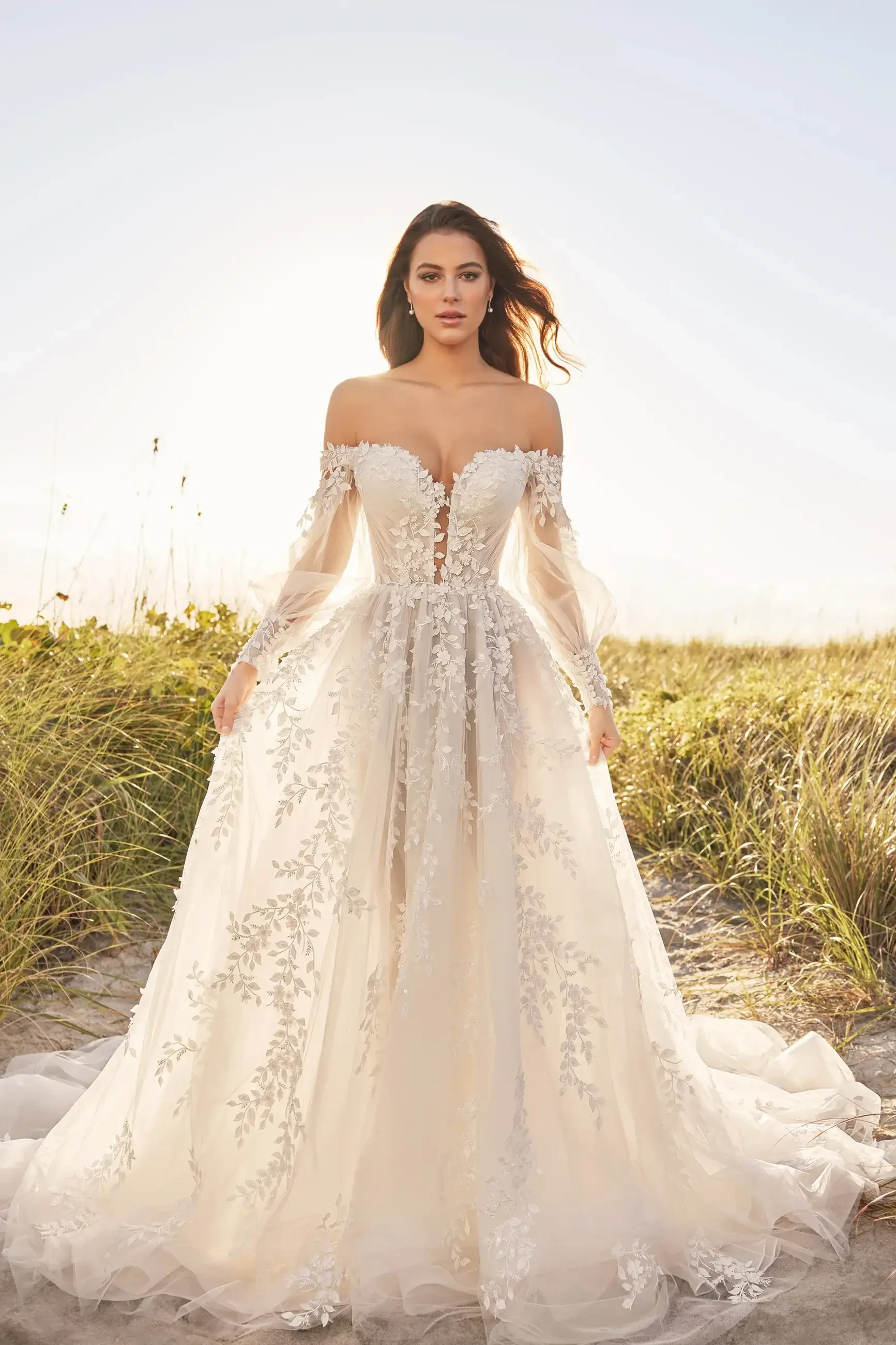 A woman in a stunning off-the-shoulder wedding dress with intricate floral embroidery stands on a sandy beach, illuminated by sunlight.