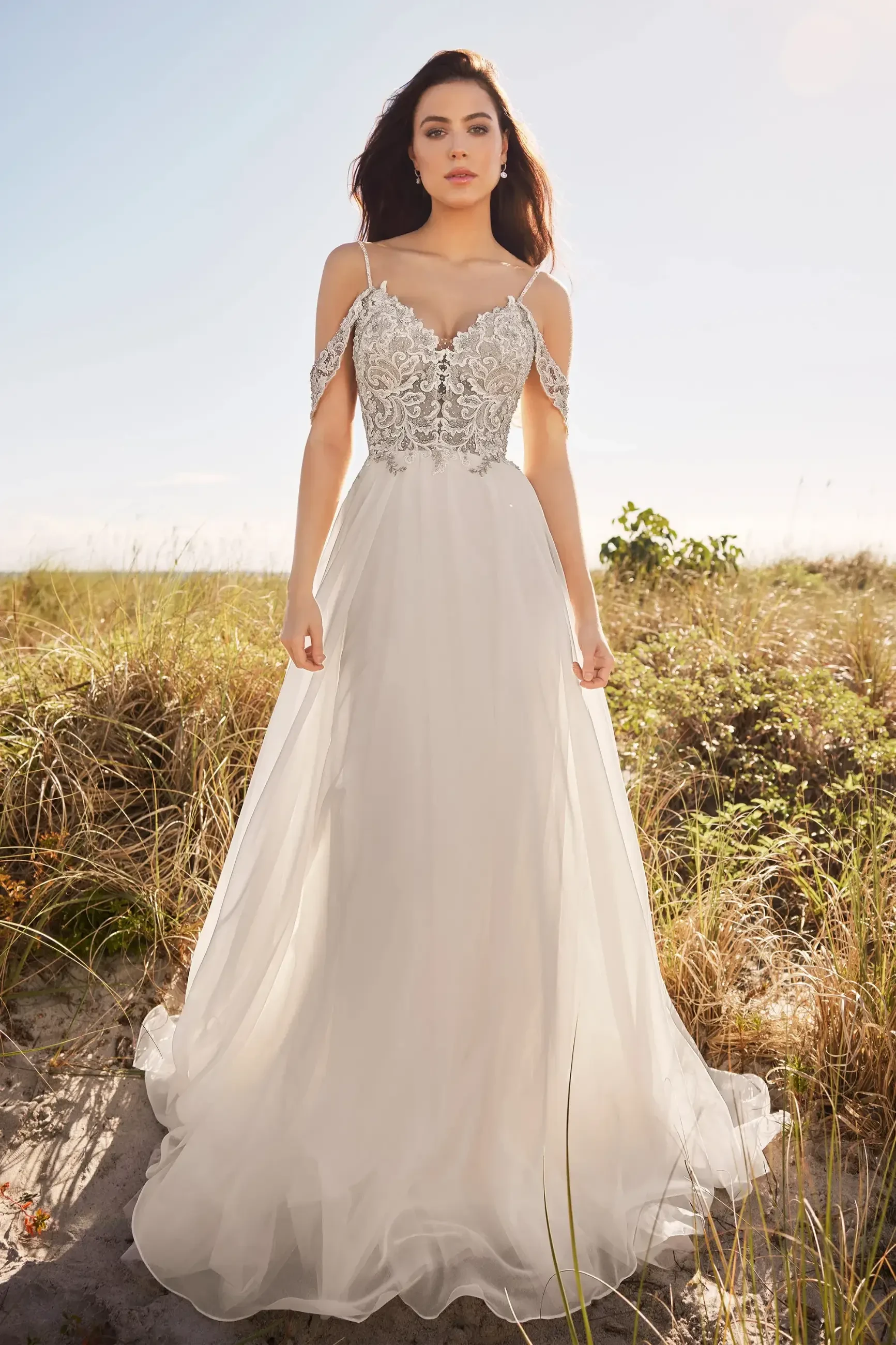 Bride in elegant white gown with intricate lace bodice and off-shoulder straps, stands confidently in sunlit grass field, evoking a serene, romantic mood.