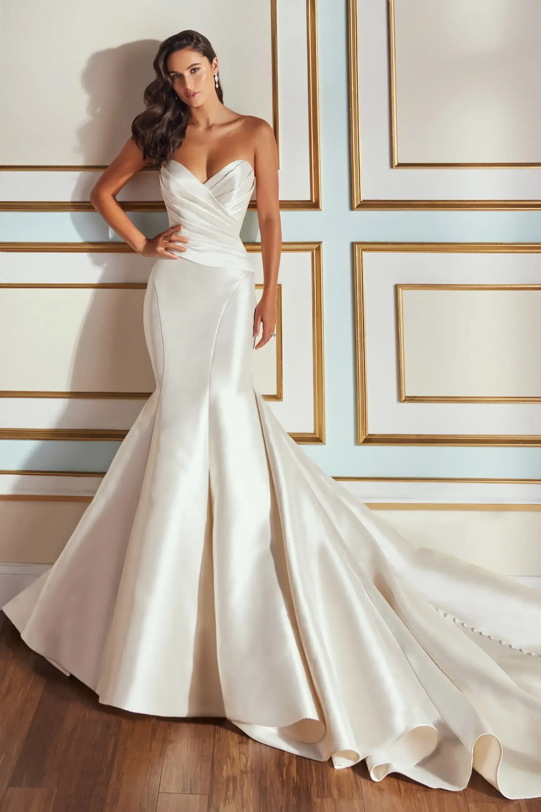 A woman in a strapless, fitted white gown with a flowing train, standing against a wall with gold trim.