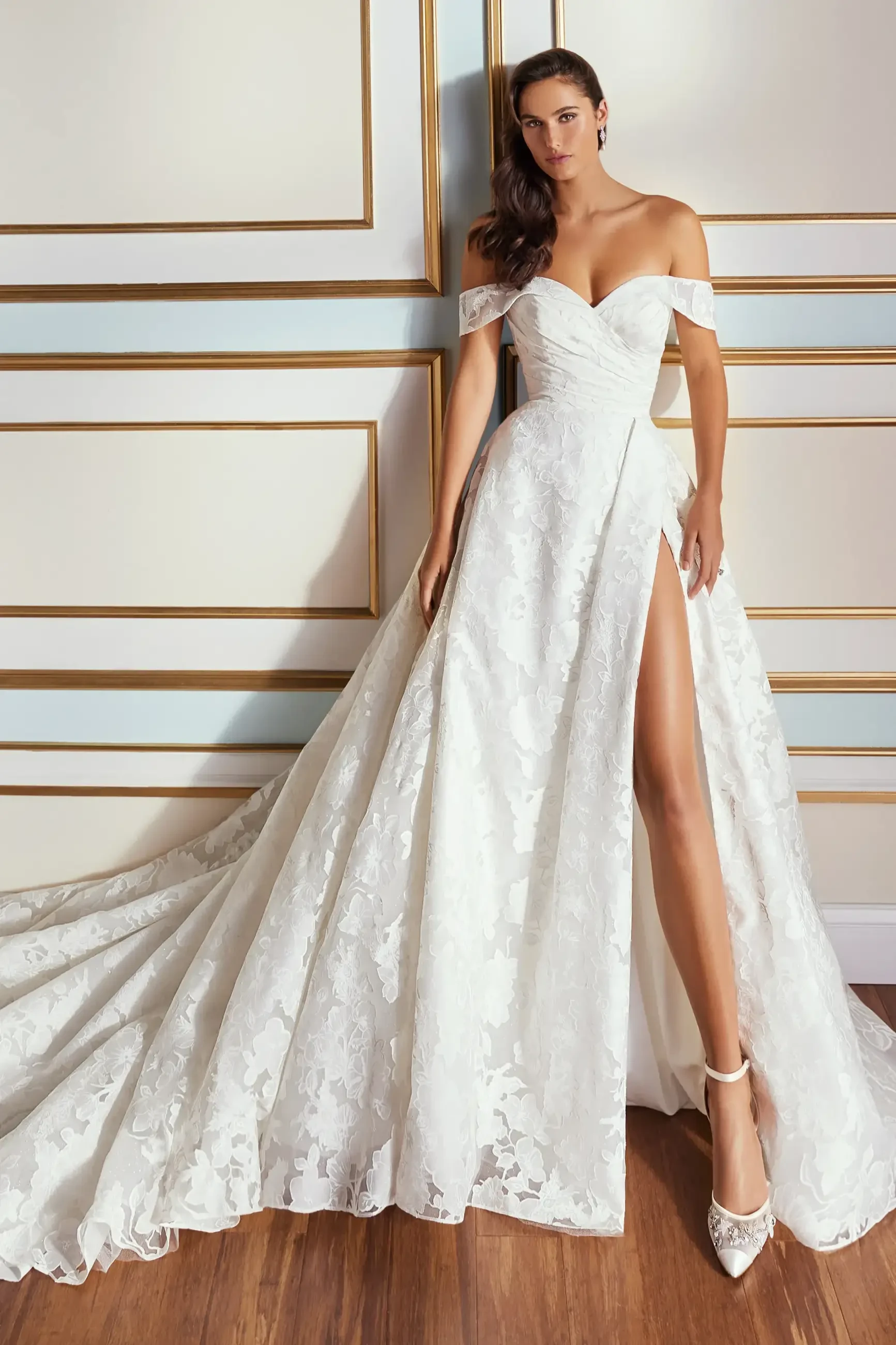 A model wearing an elegant white off-the-shoulder bridal gown with a thigh-high slit, posed against a decorative wall.