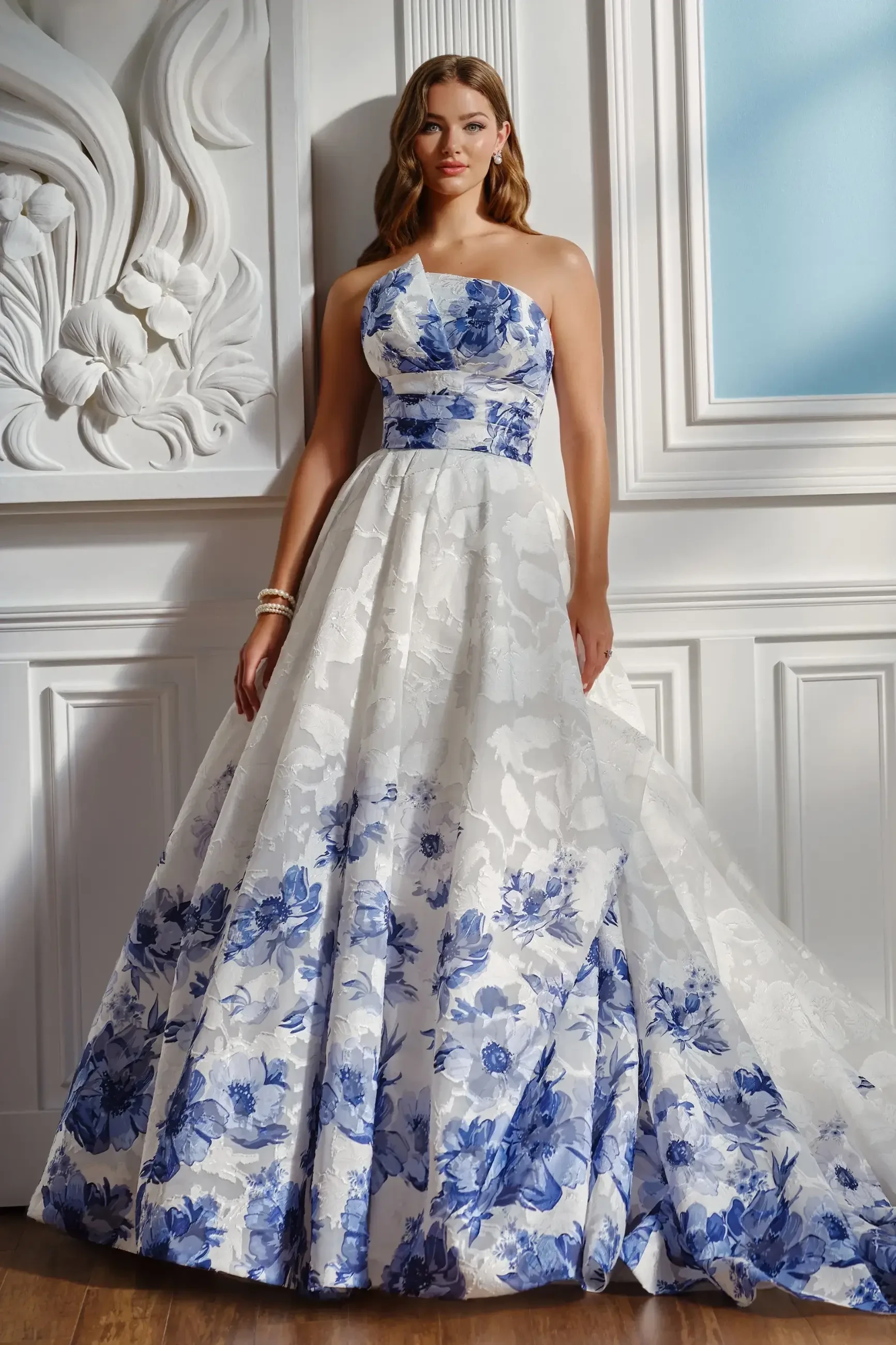A woman wearing a strapless, floral-patterned ball gown with blue and white colors, standing gracefully against a white ornate wall.