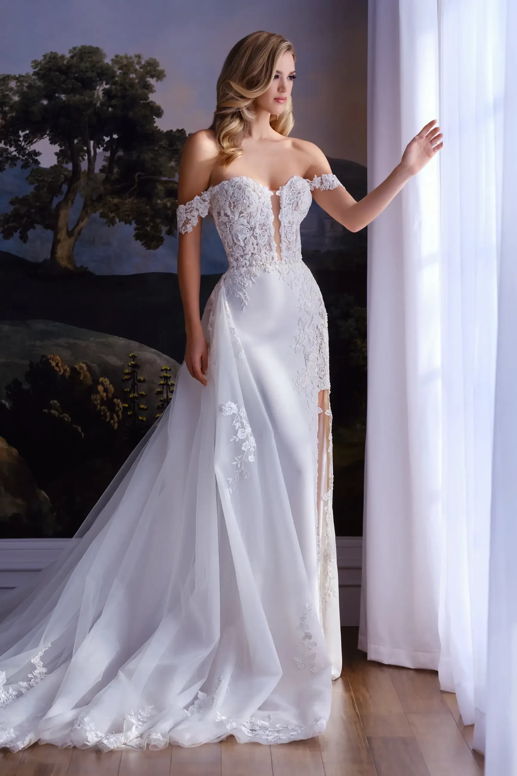 Model is wearing a crepe and lace mermaid gown with a plunging sweetheart neckline, semi-sheer bodice, removable off-the-shoulder straps, and a detachable train.